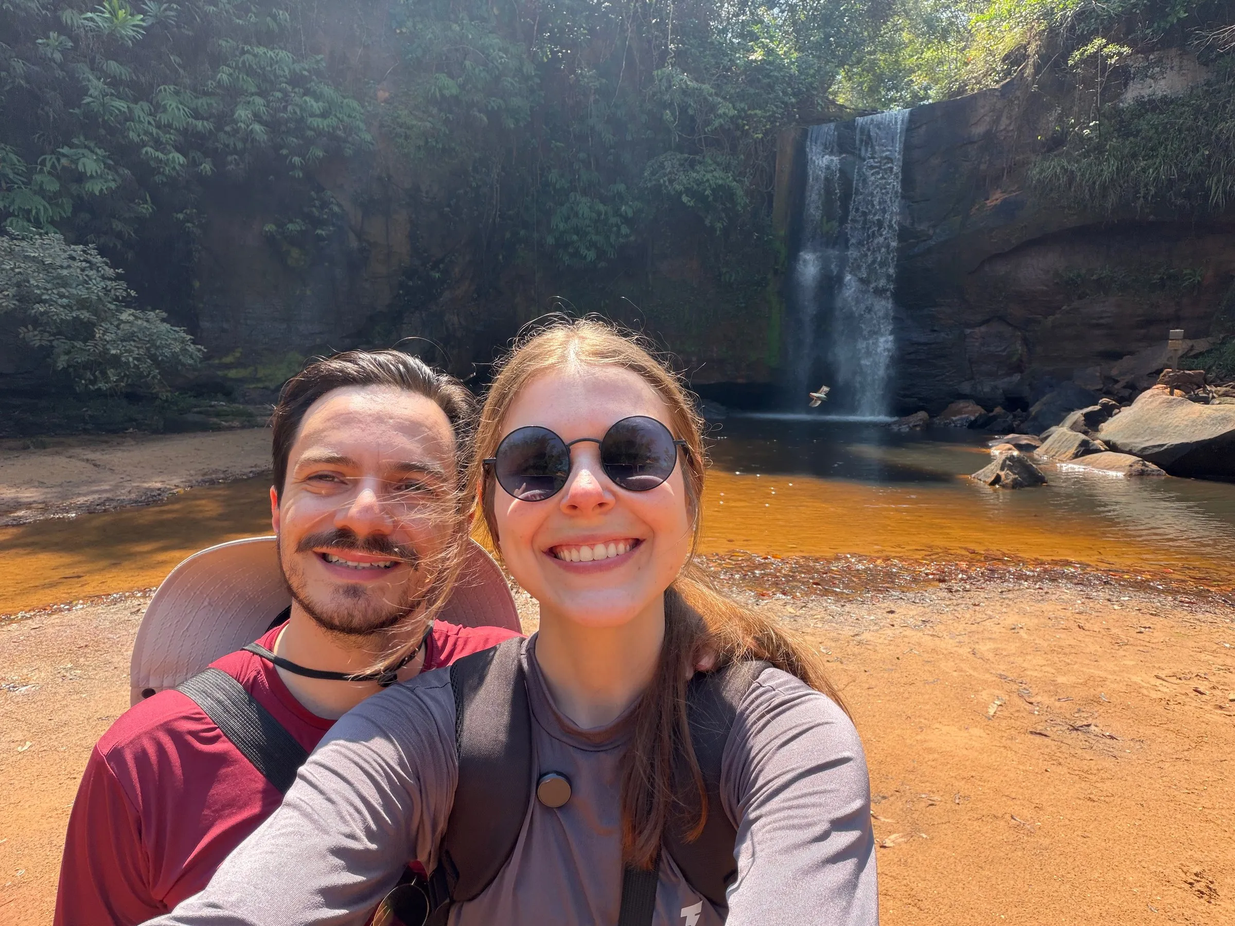 Lucas e Gabriella sorrindo numa selfie, com uma cachoeira e um poço de água ao fundo