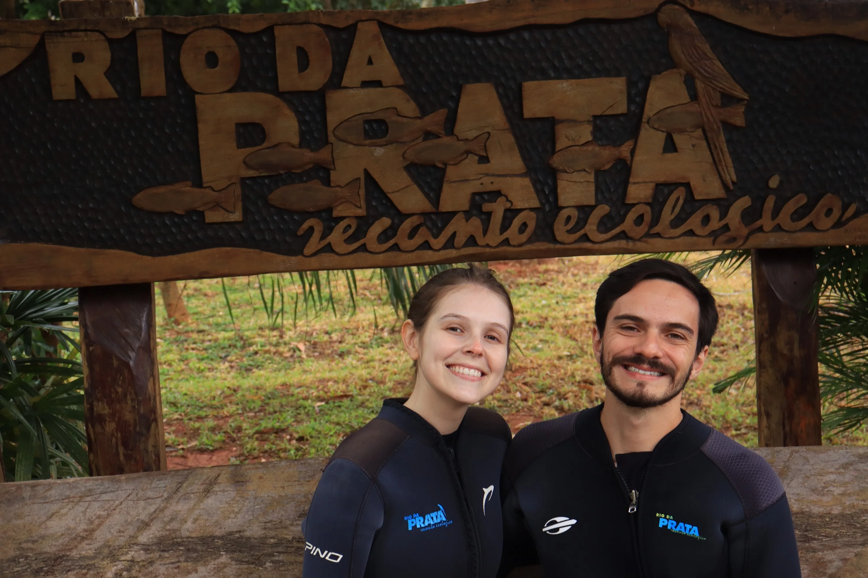 Lucas e Gabriella sorrindo de roupa de neoprene sob a placa do Recanto Ecológico Rio da Prata