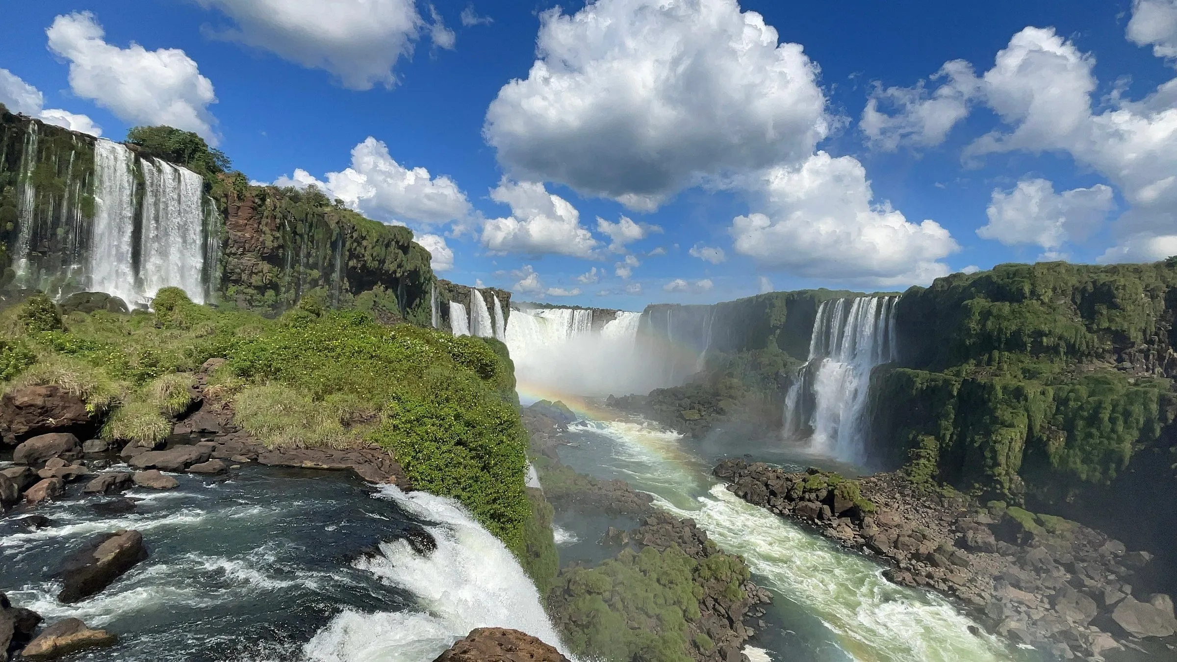 Cataratas do Iguaçu com arco-íris sobre o rio, vistas do alto, com céu azul e nuvens